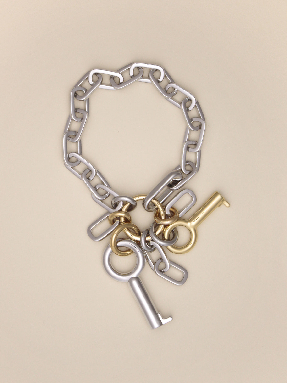 Laura Lombardi Permesso Bracelet, a silver chain bracelet with hanging keys and chains in mixed silver and gold finishes, on a neutral tan background.