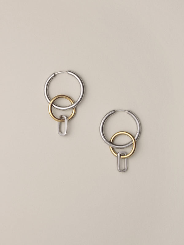 Side view of Laura Lombardi Two Tone Passa Earrings, medium silver hoops with a gold circular charm and a silver oval charm.